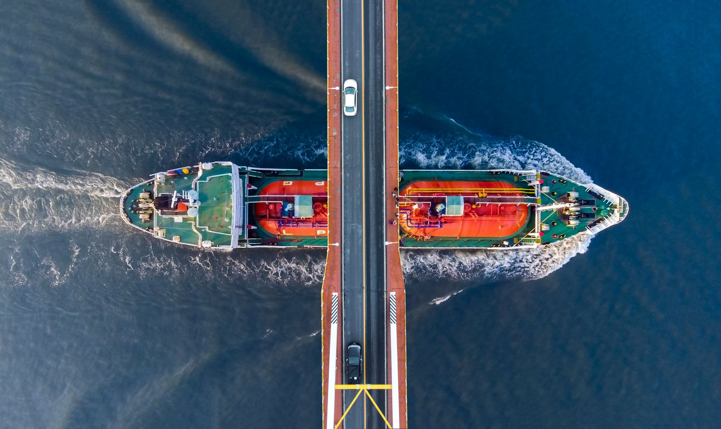Aerial view of a large cargo ship passing under a bridge with a driving car Aerial view of a large cargo ship passing under a bridge with a driving car